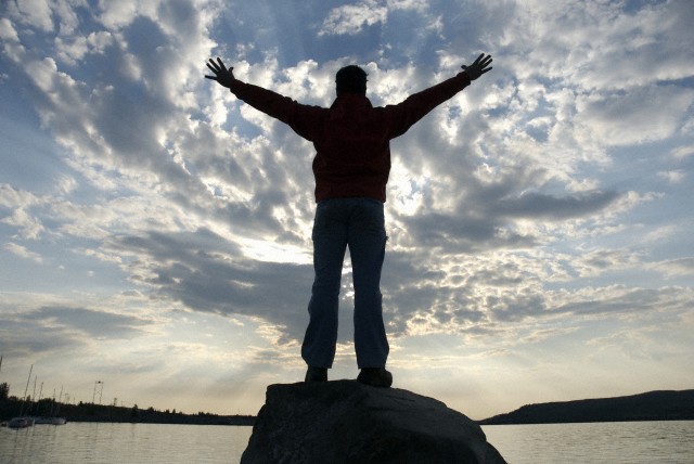 Man standing on rock near water, arms outstretched, rear view